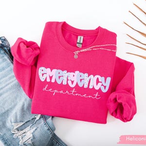May include: A hot pink sweatshirt with the words "emergency department" in a playful font. The word "emergency" is in a light blue and white pattern. A silver necklace and distressed blue jeans are next to the sweatshirt.