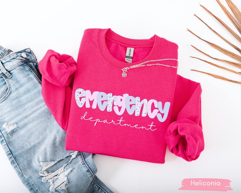 May include: A hot pink sweatshirt with the words "emergency department" in a playful font. The word "emergency" is in a light blue and white pattern. A silver necklace and distressed blue jeans are next to the sweatshirt.