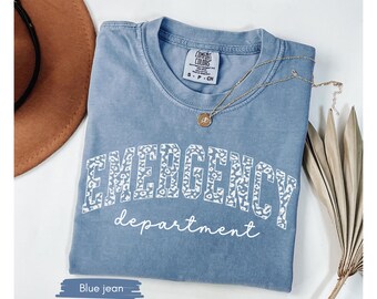 May include: A blue jean colored t-shirt with the words "EMERGENCY department" printed on the front in white. The word "EMERGENCY" has a leopard print design. A gold necklace and a brown hat are also in the image.