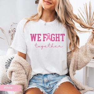 K&ouml;nnte beinhalten: Wei&szlig;es T-Shirt mit dem pinkfarbenen Text "WE FIGHT together" und einer pinkfarbenen Schleife. Das Shirt wird mit Jeansshorts und einer beigen Strickjacke kombiniert. Das Bild wurde in einem gut beleuchteten Innenraum aufgenommen.