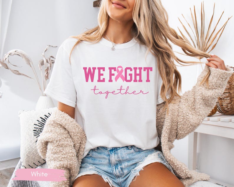 K&ouml;nnte beinhalten: Wei&szlig;es T-Shirt mit dem pinkfarbenen Text "WE FIGHT together" und einer pinkfarbenen Schleife. Das Shirt wird mit Jeansshorts und einer beigen Strickjacke kombiniert. Das Bild wurde in einem gut beleuchteten Innenraum aufgenommen.