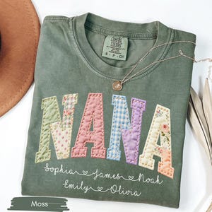 May include: A moss green t-shirt with the word "NANA" in patchwork letters. The letters are in various floral and patterned fabrics. Below the word are names and hearts. A gold necklace and a brown hat are also in the image.