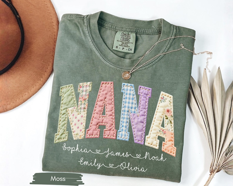 May include: A moss green t-shirt with the word "NANA" in patchwork letters. The letters are in various floral and patterned fabrics. Below the word are names and hearts. A gold necklace and a brown hat are also in the image.