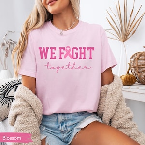 K&ouml;nnte beinhalten: Hellrosa T-Shirt mit dem Schriftzug "WE FIGHT together" in Rosa und einer rosa Schleife. Das Shirt ist im l&auml;ssigen Stil und aus weichem Material.