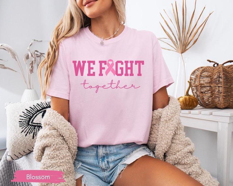 K&ouml;nnte beinhalten: Hellrosa T-Shirt mit dem Schriftzug "WE FIGHT together" in Rosa und einer rosa Schleife. Das Shirt ist im l&auml;ssigen Stil und aus weichem Material.