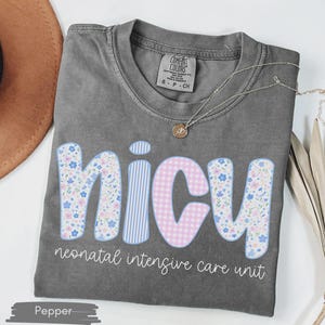 Peut inclure: Un t-shirt gris avec le mot "NICU" dans un design floral, ray&eacute; et &agrave; carreaux. Le texte "neonatal intensive care unit" est en dessous. Un collier dor&eacute; et un chapeau marron sont &eacute;galement visibles.
