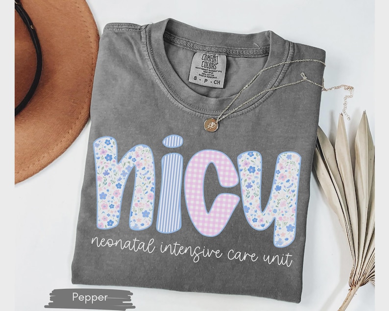 Peut inclure: Un t-shirt gris avec le mot "NICU" dans un design floral, ray&eacute; et &agrave; carreaux. Le texte "neonatal intensive care unit" est en dessous. Un collier dor&eacute; et un chapeau marron sont &eacute;galement visibles.