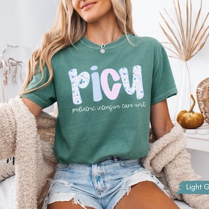 Puede incluir: Camiseta verde claro con las letras "PICU" en un dise&ntilde;o floral, a rayas y de cuadros. Las palabras "pediatric intensive care unit" est&aacute;n impresas debajo. La camiseta es de manga corta y cuello redondo.