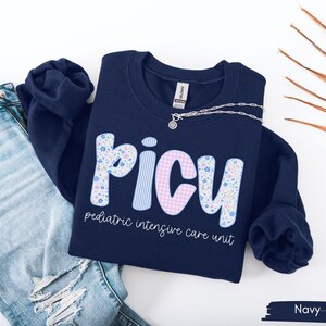 May include: Navy blue sweatshirt with the letters "PICU" in a floral, striped, and gingham pattern. The words "pediatric intensive care unit" are printed below the letters. A silver necklace is draped on the sweatshirt. The sweatshirt is next to a pair of blue jeans.