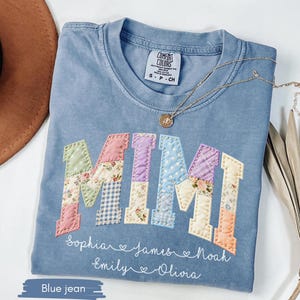 May include: A blue jean Comfort Colors t-shirt with the word "MIMI" appliqued in colorful patchwork fabric. Below the word are the names Sophia, James, Noah, Emily, and Olivia. A gold necklace rests on the shirt.