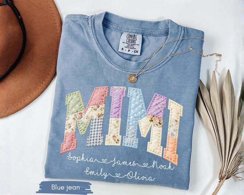 May include: A blue jean Comfort Colors t-shirt with the word "MIMI" appliqued in colorful patchwork fabric. Below the word are the names Sophia, James, Noah, Emily, and Olivia. A gold necklace rests on the shirt.