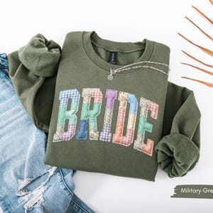 May include: An olive green sweatshirt with the word "BRIDE" appliqued in a patchwork design. The letters are in various pastel colors and patterns. A silver necklace is draped over the sweatshirt. The sweatshirt is paired with blue jeans.