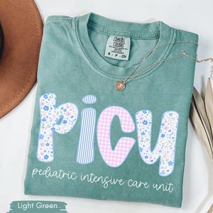 Puede incluir: Camiseta verde claro con la palabra "PICU" en un dise&ntilde;o floral, de cuadros y a rayas. El texto "pediatric intensive care unit" est&aacute; debajo de la palabra PICU. Un collar dorado y un sombrero marr&oacute;n est&aacute;n en el fondo.
