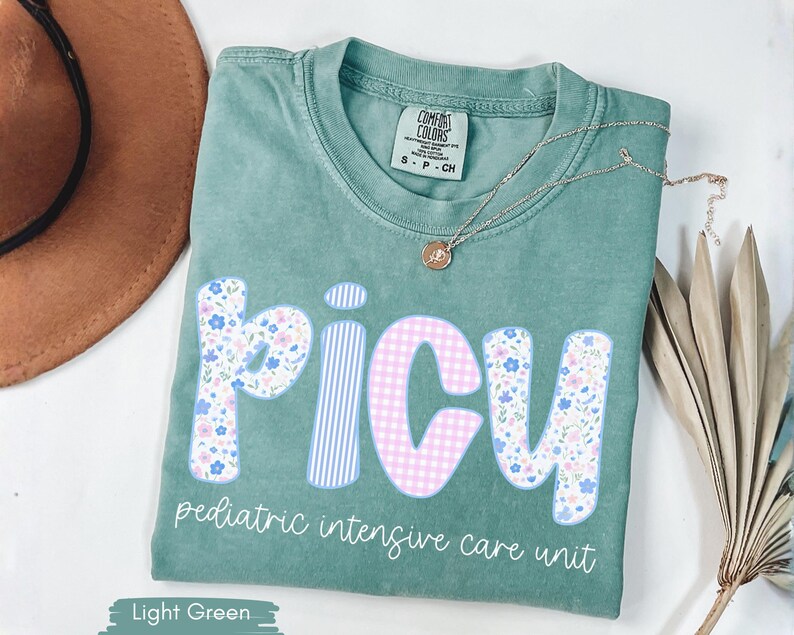 Puede incluir: Camiseta verde claro con la palabra "PICU" en un dise&ntilde;o floral, de cuadros y a rayas. El texto "pediatric intensive care unit" est&aacute; debajo de la palabra PICU. Un collar dorado y un sombrero marr&oacute;n est&aacute;n en el fondo.