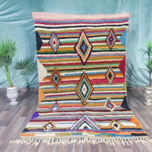 Może przedstawiać: Kolorowy, ręcznie robiony dywan z geometrycznym wzorem rombów. Dywan posiada poziome paski w różnych kolorach, w tym czerwonym, pomarańczowym, zielonym i niebieskim. Kształty rombów są w kontrastujących kolorach. Dywan ma frędzelkową krawędź.