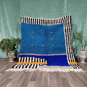 Op de afbeelding: Een levendig blauw en zwart Marokkaans tapijt met een geometrisch ontwerp. Het tapijt heeft een centraal blauw veld met gele accenten, omzoomd door zwart-witte strepen. Het tapijt wordt op een houten vloer getoond.