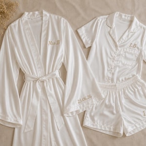 May include: A white satin robe with wide sleeves and a matching short-sleeved button-down pyjama set. The robe features the embroidered text "Mrs B" and "25.06.26". The pyjama top and shorts are embroidered with the initials "LB" in gold thread.