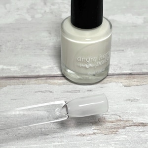 May include: A bottle of Angry Hare Polish in a light cream color with a black cap. The bottle is next to a clear plastic swatch with the same cream-colored polish applied. The bottle reads "angry hare polish" and "15 ml / .5 fl oz."