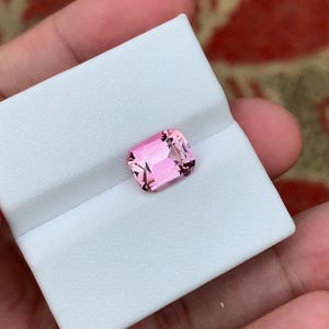 以下が含まれることがあります： 透明感のあるファセットカットが施された長方形のピンク色の宝石。鋭い幾何学的なエッジと光沢のある表面を持ち、白いテクスチャー加工が施された長方形のジュエリーディスプレイ用インサートの中に収められています。