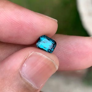 Puede incluir: Una gema rectangular facetada de color turquesa vibrante. La gema se sostiene entre los dedos, mostrando sus superficies pulidas y reflejos internos. La forma de la gema es un rectángulo con esquinas cortadas, lo que sugiere una alta calidad.