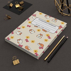 Op de afbeelding: Een crèmekleurig notitieboek met een herhalend patroon van cartoonhamsters, gele bloemen en roze strikjes. Het witte label op de kaft bevat de tekst Composition Notebook, 150 pages, Ruled, 5,71 x 8,07 inches. Gouden en zwarte potloden liggen ernaast.