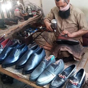 May include: A shoemaker works on a pair of brown leather shoes.  He is using a tool to polish the shoes.  There are many other shoes in the background, all in various shades of blue and brown.
