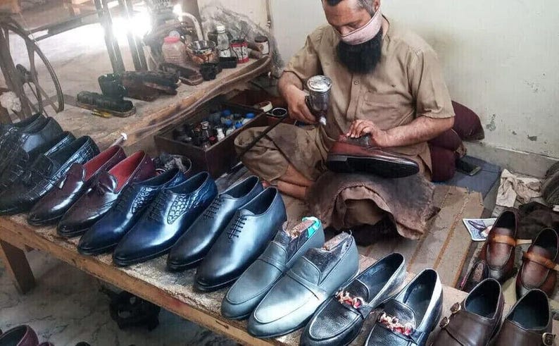 May include: A shoemaker works on a pair of brown leather shoes.  He is using a tool to polish the shoes.  There are many other shoes in the background, all in various shades of blue and brown.