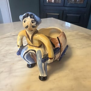 May include: A ceramic planter in the shape of a person pushing a barrel. The figure wears a yellow jacket, blue hat, and pants with blue and white stripes. The barrel is brown and tan with blue accents.