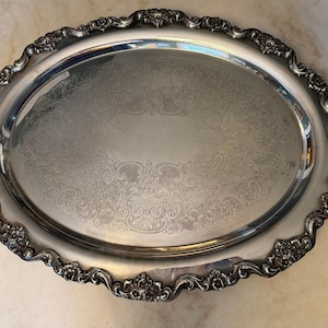 Puede incluir: Una bandeja de servicio ovalada de tono plateado con un borde ornamentado en relieve de motivos florales y volutas. La superficie central de la bandeja muestra un delicado patrón grabado de adornos simétricos, junto a dos asas integradas.