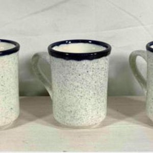 May include: Three ceramic mugs with a speckled off-white finish and a solid dark blue rim. Each mug features a cylindrical body, a curved handle on the side, and a smooth texture. The mugs are arranged in a horizontal row, showing consistent design.