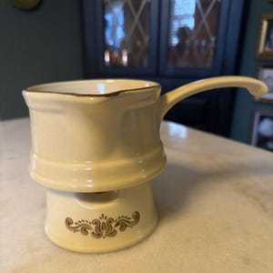 May include: Cream-colored ceramic sauce warmer with a handle and a decorative brown floral design. The warmer has a separate base with a small opening. The rim of the sauce warmer is trimmed with brown.