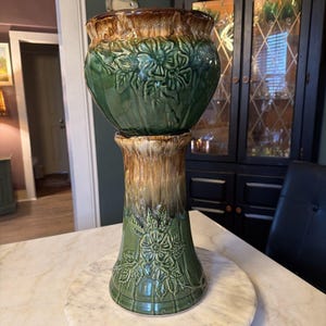 May include: A two-piece ceramic planter in shades of green and brown. The top pot features embossed floral designs and a glossy finish. The pedestal base has similar floral detailing and a textured, matte finish. The planter sits on a marble surface.