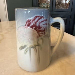 May include: A ceramic pitcher with a handle, featuring a floral design. The pitcher has a light blue gradient at the top, transitioning to white. The flowers are a mix of red and white, with green stems and leaves. The pitcher is likely for decorative use.