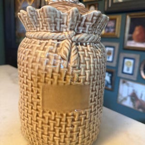 Op de afbeelding: Een lichtbruine keramische voorraadpot met een getextureerd oppervlak dat lijkt op een geweven jute zak. De pot heeft een sculpturaal touwdetail rond de hals en een bijpassend deksel met een handvat in touwstijl. Een glad rechthoekig paneel is gecentreerd.