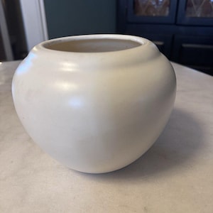 May include: A cream-colored, round ceramic vase with a wide opening. The vase has a smooth, matte finish and sits on a marble surface. The vase is a decorative item.