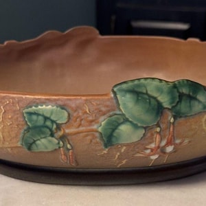 May include: A brown ceramic bowl with a scalloped rim and handles. The bowl features green leaf and flower designs. It sits on a dark brown base. The bowl is likely for decorative purposes.