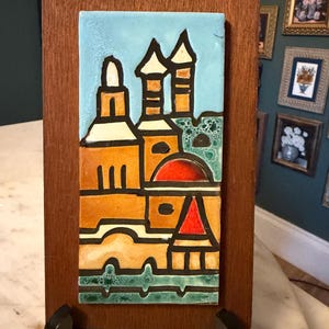 May include: A rectangular ceramic tile mounted on a dark wood plaque, featuring a stylized architectural scene with a light blue sky, orange buildings, a red dome, a red roof, and green accents, all outlined in thick black lines on a white background.