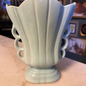 May include: A pale blue ceramic vase featuring a scalloped, fan-shaped top edge and vertical ribbed detailing. The sides of the vase are adorned with a series of open, circular cutouts, and the piece rests on a sturdy, stepped pedestal base.