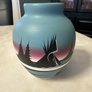 May include: A hand-painted ceramic vase with a teal blue background. The vase features a landscape design with black trees, a teepee, and a white and red sunset. A white stripe accents the base.