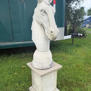 May include: A light gray stone sculpture of a horse head bust mounted on a matching square pedestal. The pedestal features carved horse head reliefs on its sides. The sculpture has a weathered texture and is positioned in an upright vertical orientation.