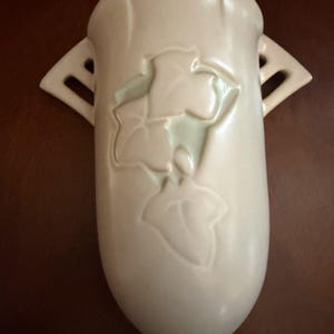 May include: Cream-colored ceramic wall vase with a leaf design. The vase has a rounded bottom and two side handles with cut-out details. The leaf design is slightly raised and lighter in color.
