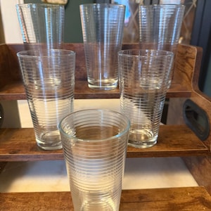 May include: A set of six clear glass tumblers, each featuring a series of thin, horizontal etched lines encircling the exterior. The glasses are arranged in a tiered formation, with five glasses positioned on wooden shelves and one in the front.