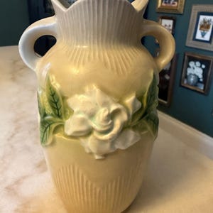 May include: A pale yellow ceramic vase featuring two side handles and a scalloped rim. The body of the vase is decorated with a raised, three-dimensional white flower blossom and green leaves, complemented by vertical ribbed detailing at the base.