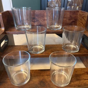 May include: A set of six clear glass tumblers arranged on a three-tiered wooden display stand. Each glass features a series of thin, horizontal etched lines circling the exterior surface, creating a subtle textured pattern on the transparent glass.