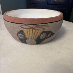 May include: A decorative ceramic bowl with a sand-colored exterior and a white interior. The rim is a reddish-brown color, with a black band below. The bowl features a stylized design of a creature with blue and yellow accents.