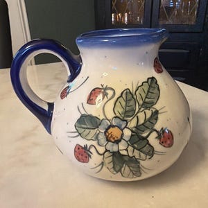 May include: A white ceramic pitcher with a blue handle and rim, decorated with red strawberries and green leaves. The pitcher has a rounded body and a spout for pouring. A charming addition to any kitchen.