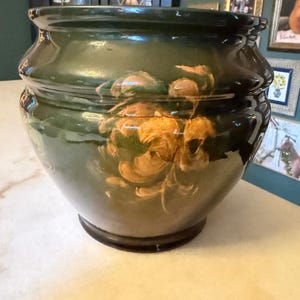 Pode incluir: Um vaso de cerâmica verde-escuro com acabamento brilhante e formato arredondado e bojudo. O exterior apresenta um design floral pintado à mão em tons de laranja e dourado, retratando um grupo de flores centrado na superfície frontal.