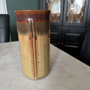 May include: A tall, handmade ceramic vase with a cylindrical shape. The top is a dark brown, transitioning to a lighter brown and then a pale yellow. Vertical lines and drips add texture. A unique piece for home decor.