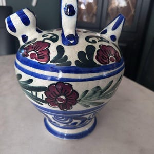 May include: A hand-painted ceramic pitcher with a white base, adorned with blue stripes and floral designs in burgundy and green. The pitcher features a handle and two spouts, showcasing a traditional, decorative style.