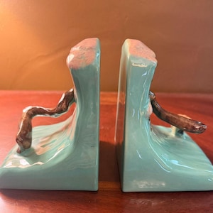 May include: A pair of ceramic bookends featuring a glossy light blue wave design with a brown, textured branch-like element extending from the center. The top edges of the wave forms are accented with a soft pink hue, creating a smooth finish.